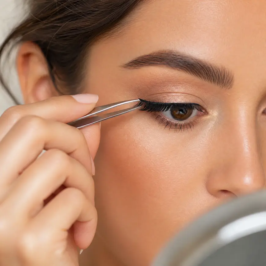 A woman carefully applying false lashes with tweezers in front of a round makeup mirror.